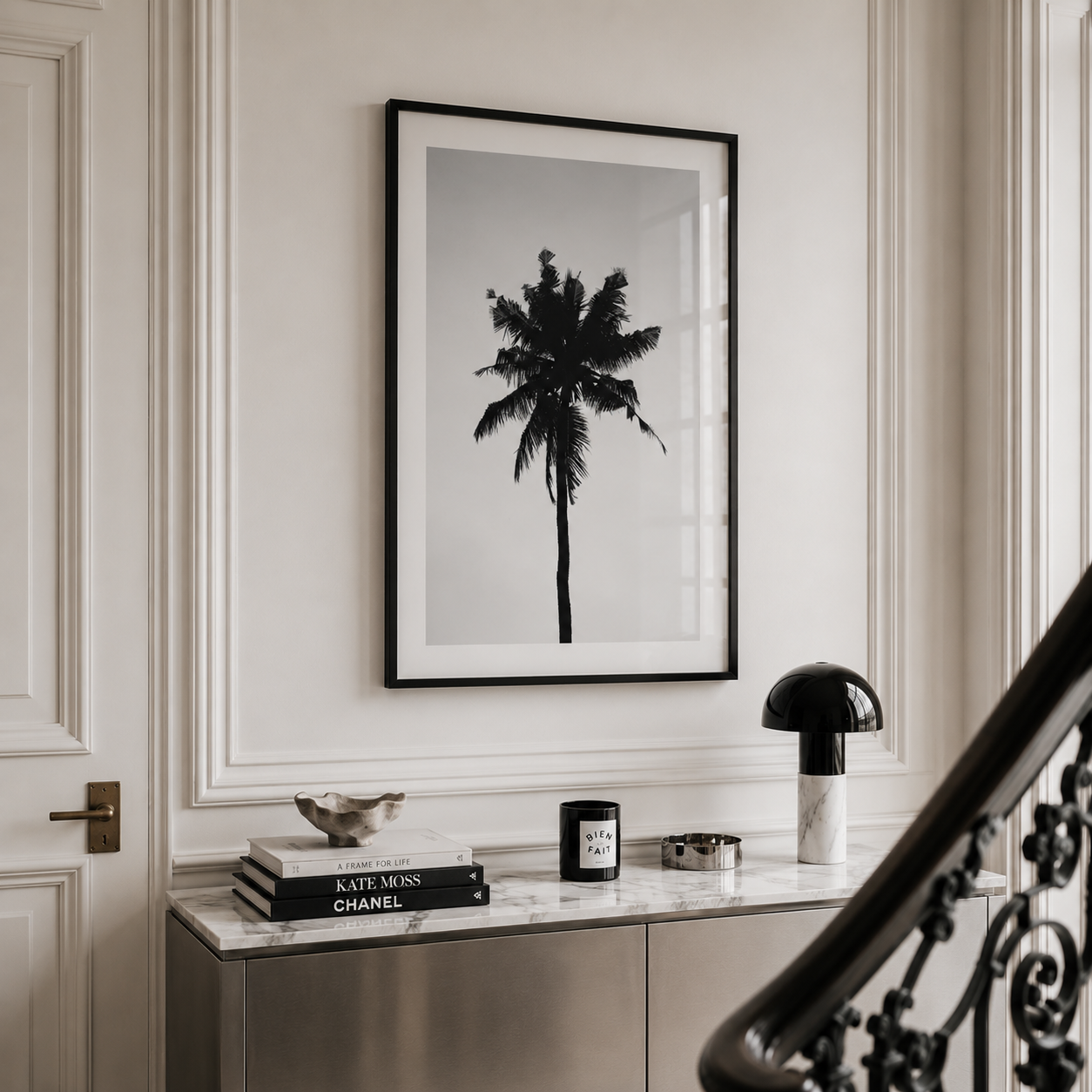 A framed black and white art print of a palm tree hangs on a white paneled wall above a marble console table styled with books, a candle, and a lamp.