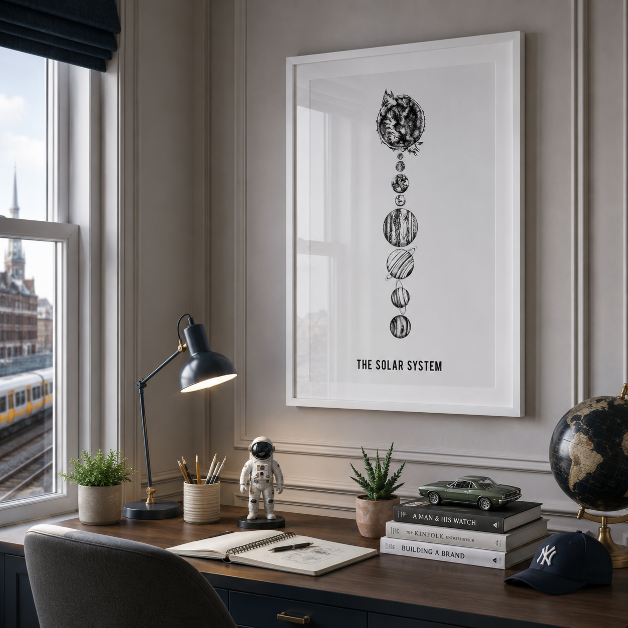 A framed black-and-white solar system print hangs on a paneled wall above a styled desk with a lamp, notebook, plants, books, and a globe in a real interior setting.