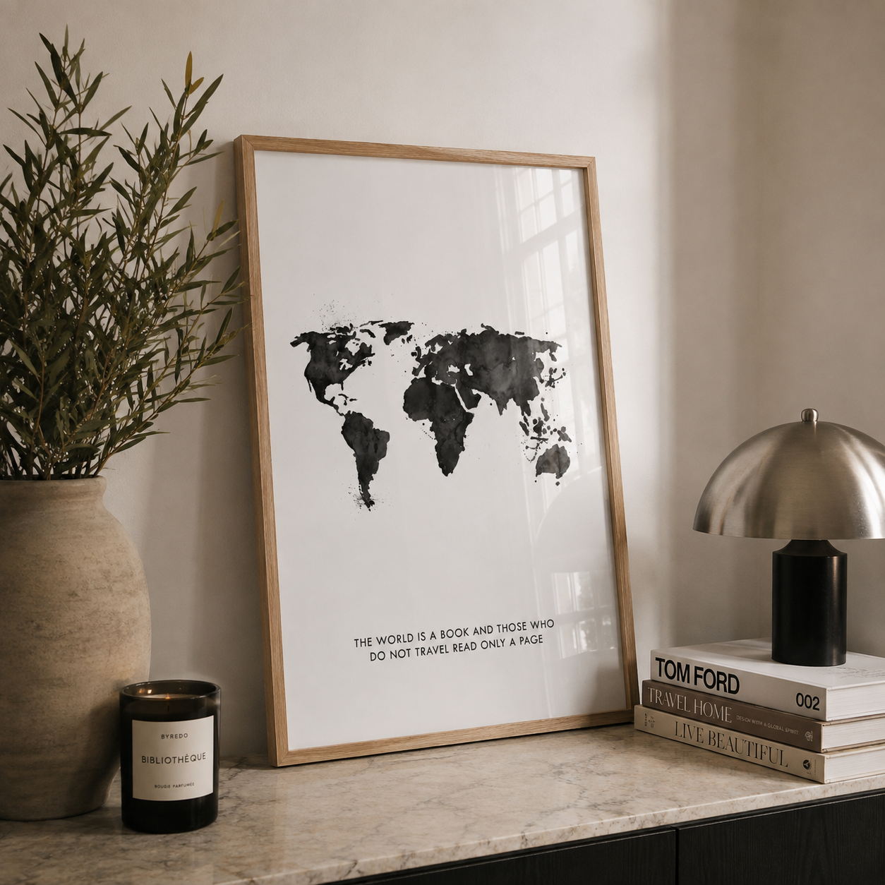 A framed world map art print with black watercolor continents on a white background, leaning on a marble console table next to a potted plant, a candle, and a stack of books with a silver dome lamp.