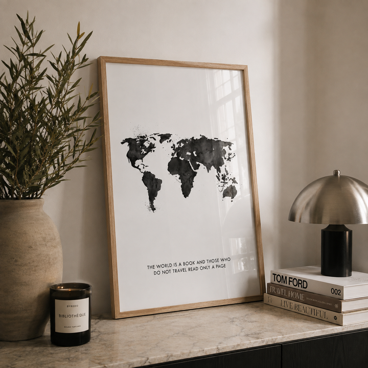 A framed world map art print with black watercolor continents on a white background, leaning on a marble console table next to a potted plant, a candle, and a stack of books with a silver dome lamp.