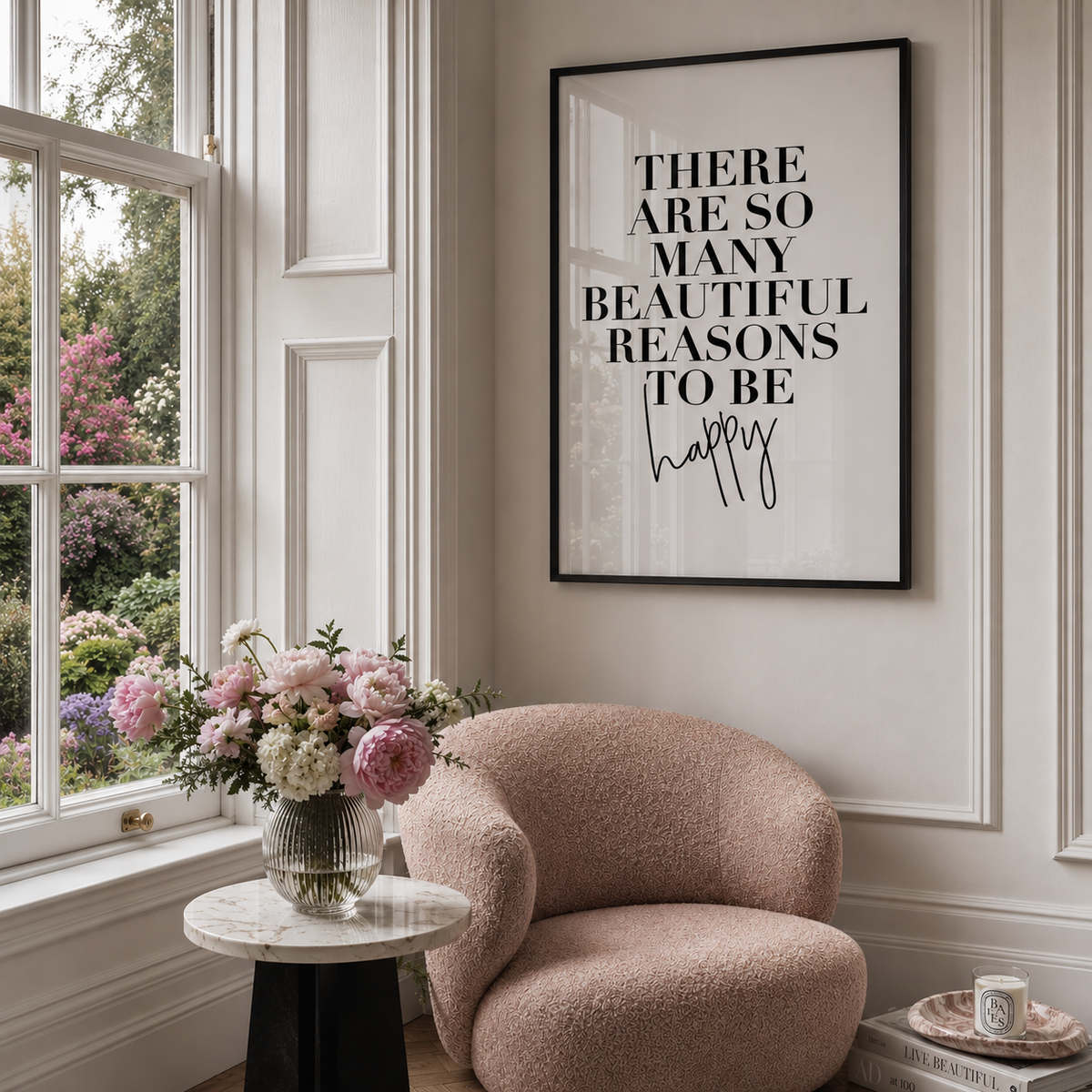 A framed typographic wall art print hangs on a cream paneled wall above a blush pink armchair, with a window, flowers, and a small table visible in a styled interior.