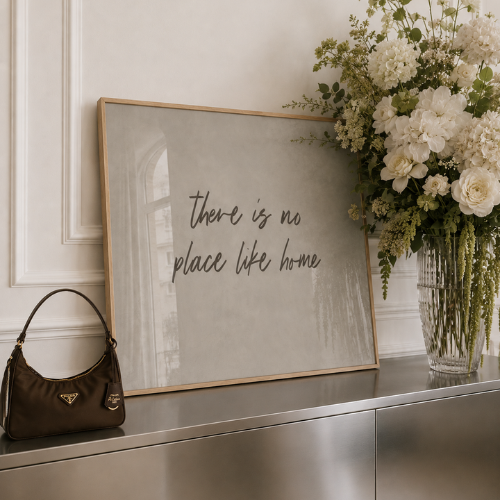 A framed wall art print with the handwritten phrase 'there is no place like home' in dark script on a light neutral background, leaning on a metallic console table beside a brown handbag and a large white floral arrangement.