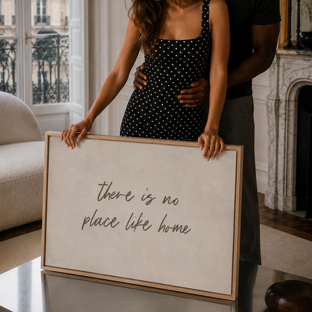 A couple in a cozy Parisian living room setting holds a large framed canvas print with the handwritten phrase 'there is no place like home'. The woman wears a Zara black and white polka dot dress, and the man wears a black t-shirt.