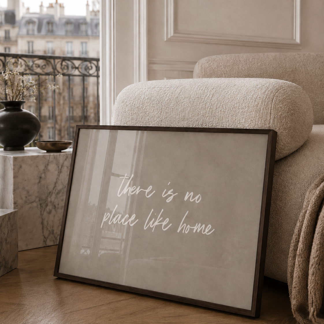 A walnut framed large wall art print leaning against a beige armchair in a luxury Parisian apartment living room. The print has a warm grey-beige background with the phrase 'there is no place like home' in white script.