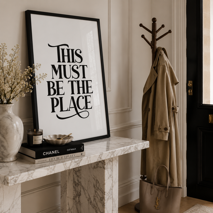 A framed typographic wall art print reading “THIS MUST BE THE PLACE” in black lettering on a white background, leaning on a marble console table in a styled interior. A tan trench coat hangs on a coat rack, with a vase, books, and a handbag nearby.