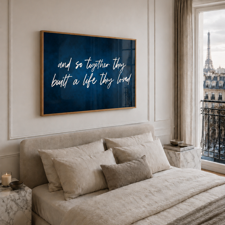 A framed wall art print with white script text on a dark blue background hangs above a neatly made bed in a softly lit Parisian bedroom. The print reads, 'and so together they built a life they loved.