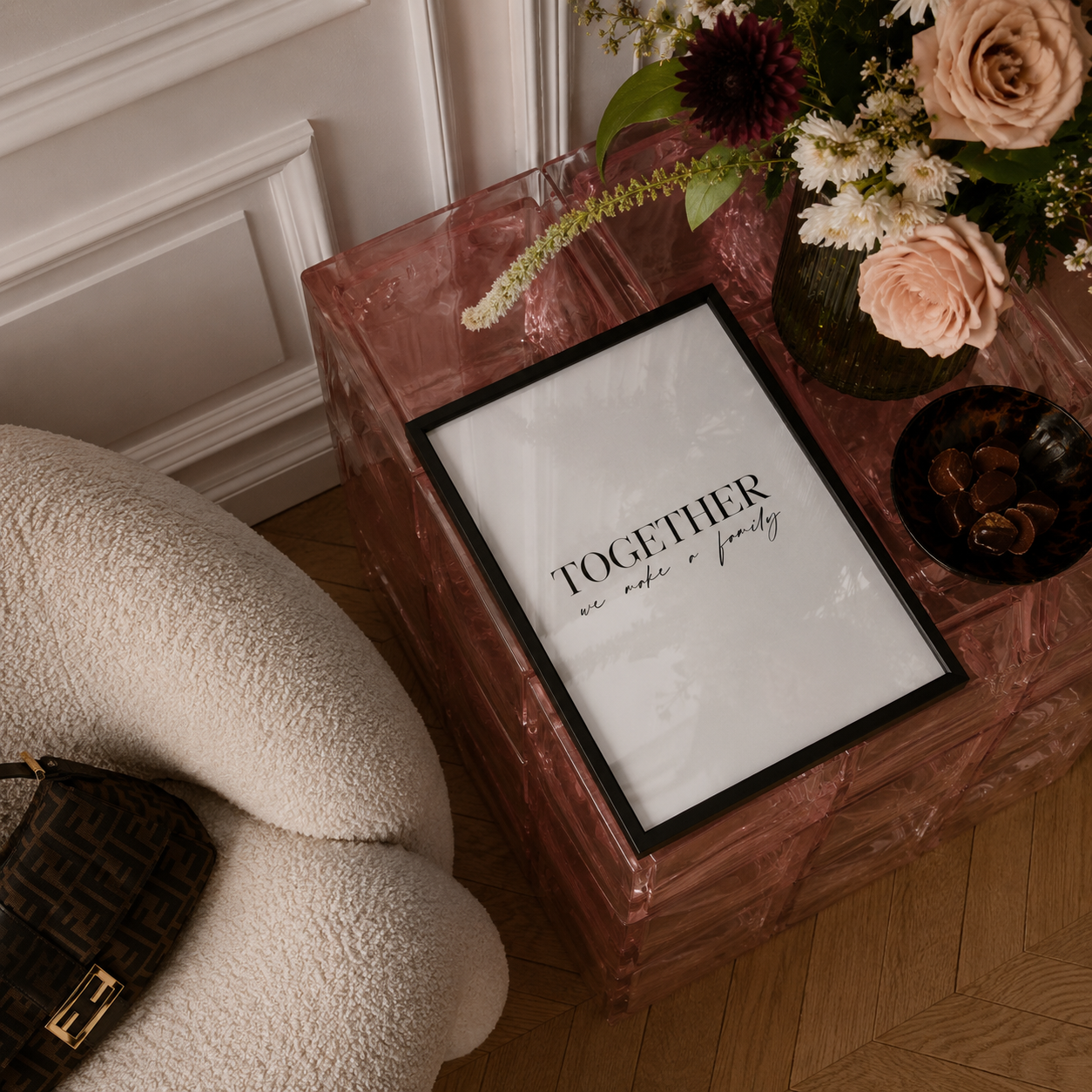 A framed wall art print with the text 'TOGETHER we make a family' in black script on a white background, displayed on a pink marble side table in a styled living room setting.