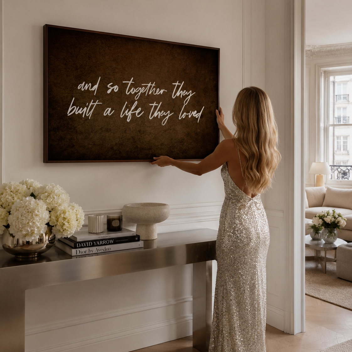 A woman in a sparkling silver gown hangs a large dark brown framed canvas print on a white wall in a bright, elegant room. The canvas features white cursive script text that reads, 'and so together they built a life they loved'.