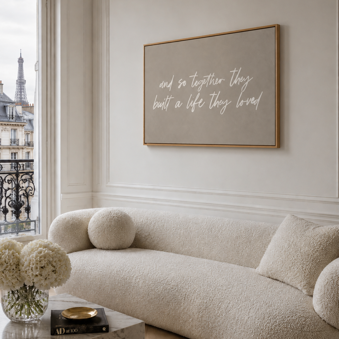 A framed large canvas wall art print with white script text reading 'and so together they built a life they loved' hangs above a cream boucle sofa in a bright, elegant room with a window and balcony view.