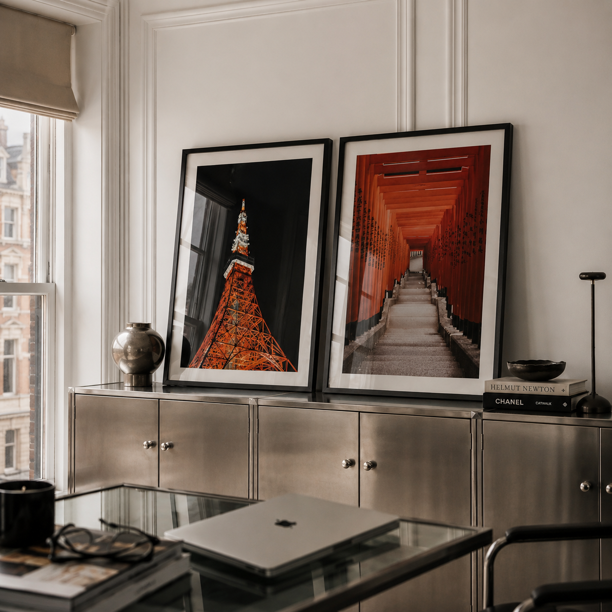 Two framed vertical art prints leaning against a white paneled wall above a metallic cabinet in a modern room. The left print shows a close-up of a red-orange pagoda against a black background, and the right print shows a red-orange corridor tunnel.