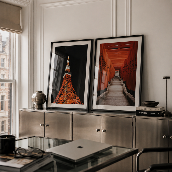 Two framed vertical art prints leaning against a white paneled wall above a metallic cabinet in a modern room. The left print shows a close-up of a red-orange pagoda against a black background, and the right print shows a red-orange corridor tunnel.