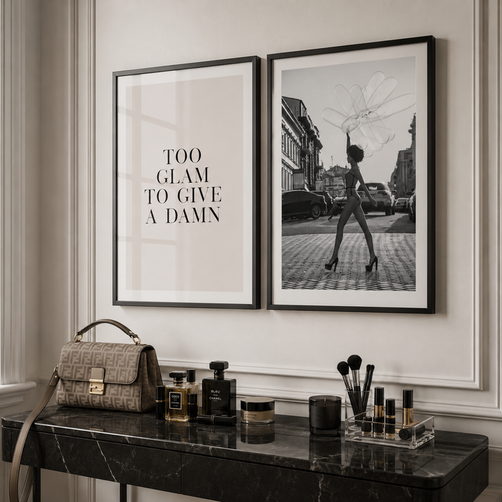 Two framed wall art prints displayed side by side above a black marble console table. The left print is a minimalist beige poster with the text 'TOO GLAM TO GIVE A DAMN' in black serif letters.