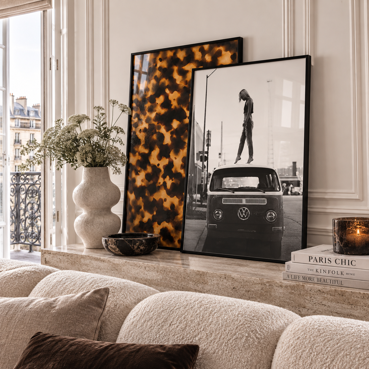 Two framed art prints leaning against a cream wall on a mantelpiece above a textured sofa. The left print features a bold black and mustard yellow abstract pattern, and the right print is a black-and-white photograph of a classic VW bus with a pedestrian crossing in front.
