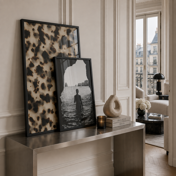 Two framed art prints leaning on a metallic console table in a stylishly decorated room. The larger print features a black and beige tortoise shell  print, and the smaller print is a black and white photograph of a figure standing in water.