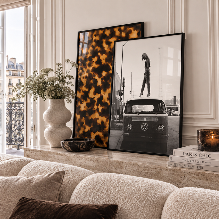 Two framed art prints leaning against a cream wall on a mantelpiece above a textured sofa. The left print features a bold black and mustard yellow abstract tortoise shell pattern, and the right print is a black-and-white photograph of a classic VW bus with a pedestrian crossing in front.
