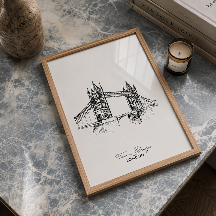 A framed black ink sketch of Tower Bridge, London, displayed on a gray marble surface with a candle, books, and a stone vase nearby.