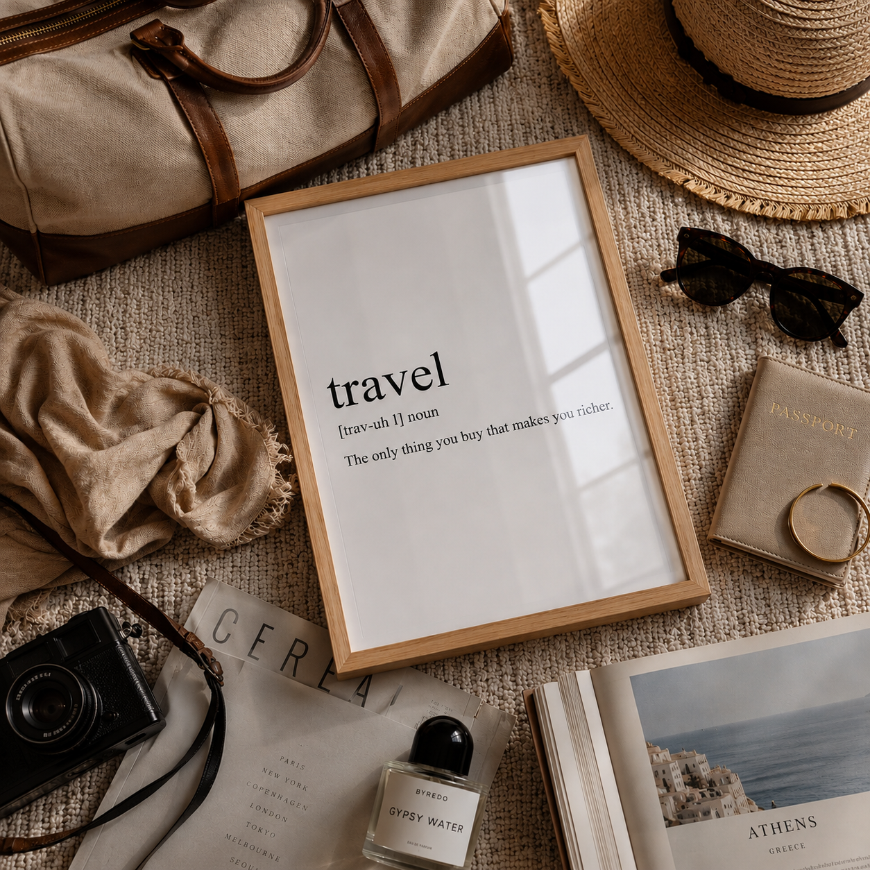 A framed typographic wall art print reading 'travel' with a dictionary-style definition, styled in a flat lay with a straw hat, sunglasses, passport, perfume bottle, camera, scarf, and books on a textured beige surface.