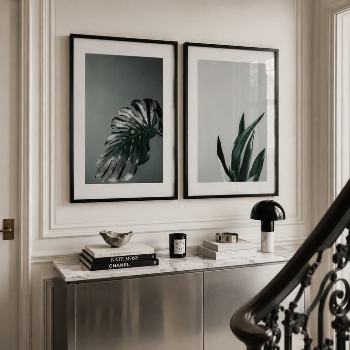 Two framed botanical art prints hanging side-by-side on a cream-colored wall above a marble console table with decorative items and a staircase railing in the foreground.