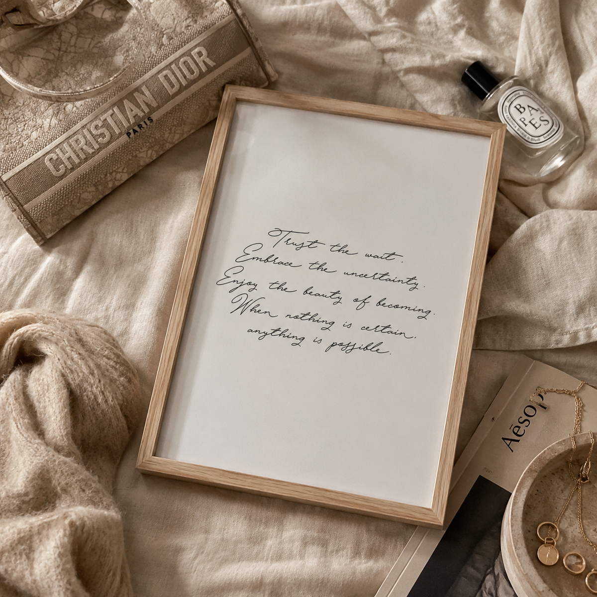 A framed typographic art print with elegant black cursive text on a white background, styled in a flat lay with a beige knitted blanket, a Dior bag, a Diptyque bottle, and gold jewelry.