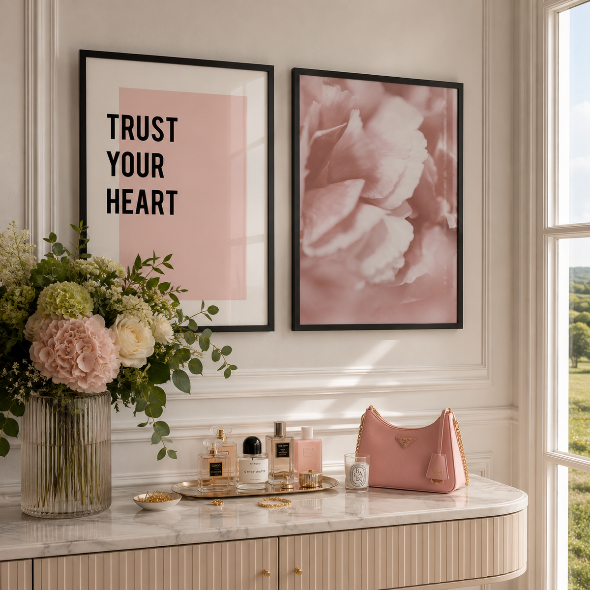 Two framed wall art prints hang above a marble console table in a bright interior. The left print reads “TRUST YOUR HEART” in black text on a blush pink background, and the right print shows a soft pink floral design.