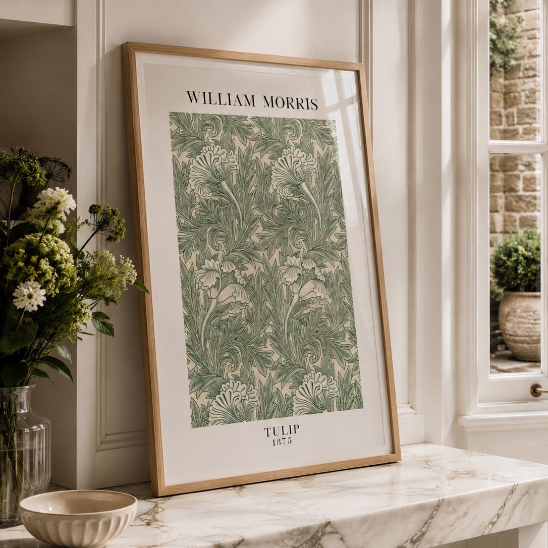 A framed William Morris Talip 1857 art print with green floral patterns on a cream background, leaning against a wall on a marble mantelpiece in a beautifully styled room.