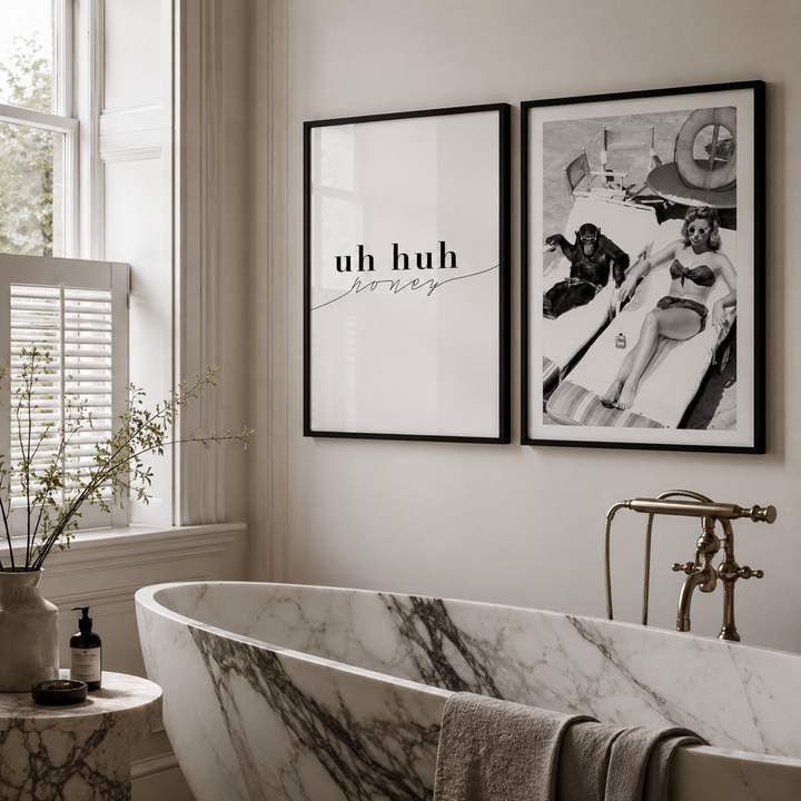 Two framed wall art prints hang above a freestanding marble bathtub in a bright bathroom. The left print is white with black text reading 'uh huh honey,' and the right print is a black-and-white retro beach scene.