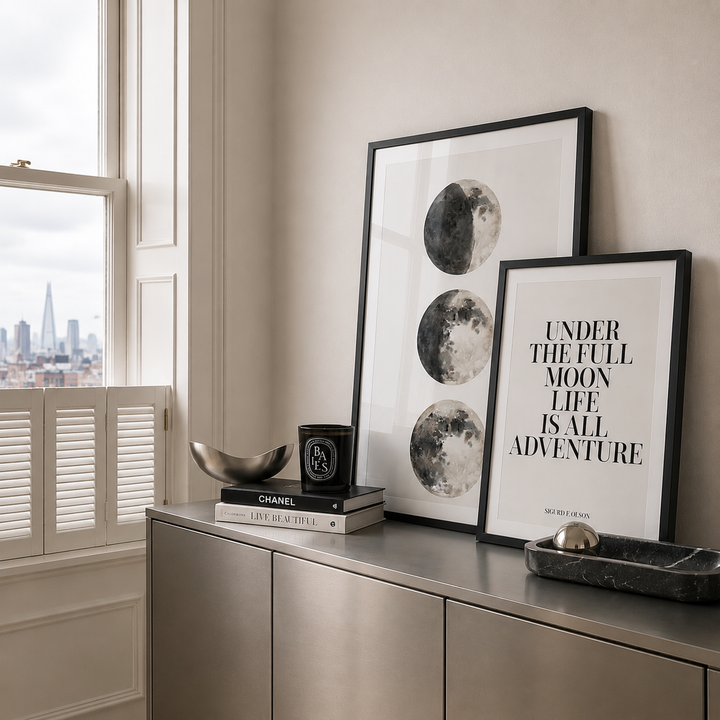 Two moon inspired framed wall art prints leaning against a beige wall on a metallic sideboard, with a window, shutters, books, and decorative objects in a styled interior setting.