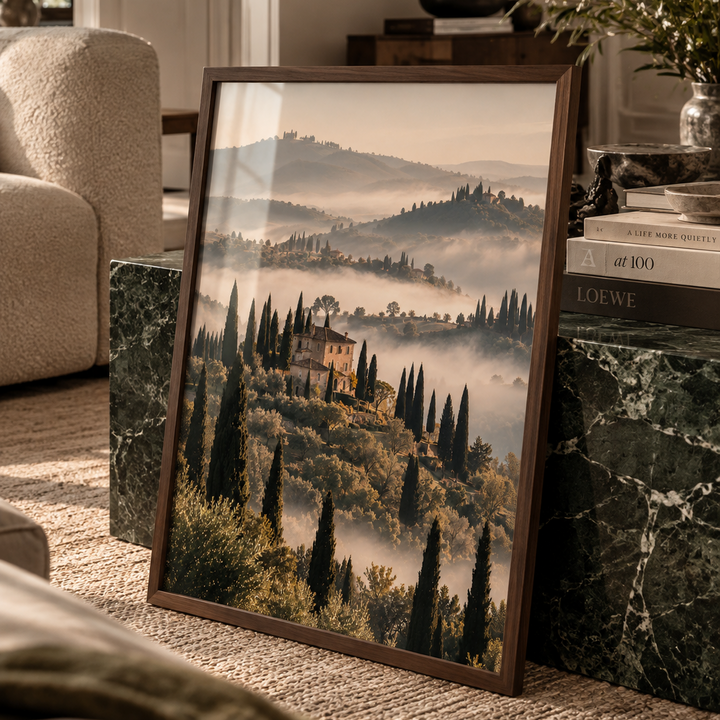 A framed landscape art print depicting a Tuscan hillside with cypress trees and a villa surrounded by misty mountains, styled leaning against a green marble side table in a cozy living room setting.