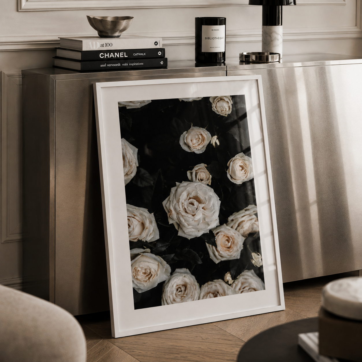 A framed floral art print featuring cream roses on a black background, leaning against a metallic cabinet in a styled interior setting.