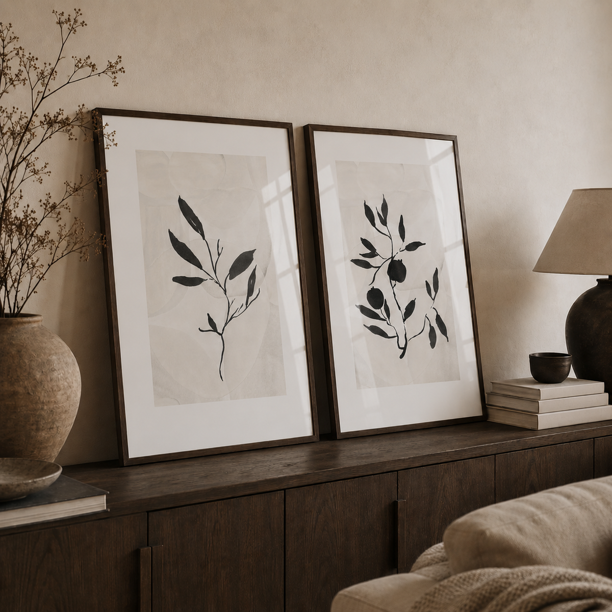 Two framed botanical art prints with black leafy illustrations on a warm off-white background, leaning on a dark wood console in a styled living room setting.
