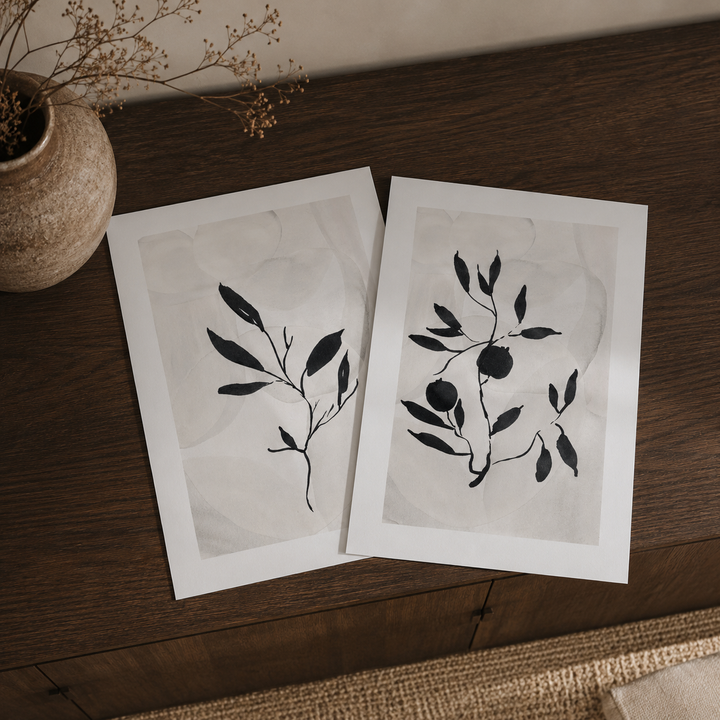 Two minimalist wabi-sabi inspired botanical art prints with black leaf illustrations on white paper, displayed on a dark wooden table next to a textured vase with dried branches.