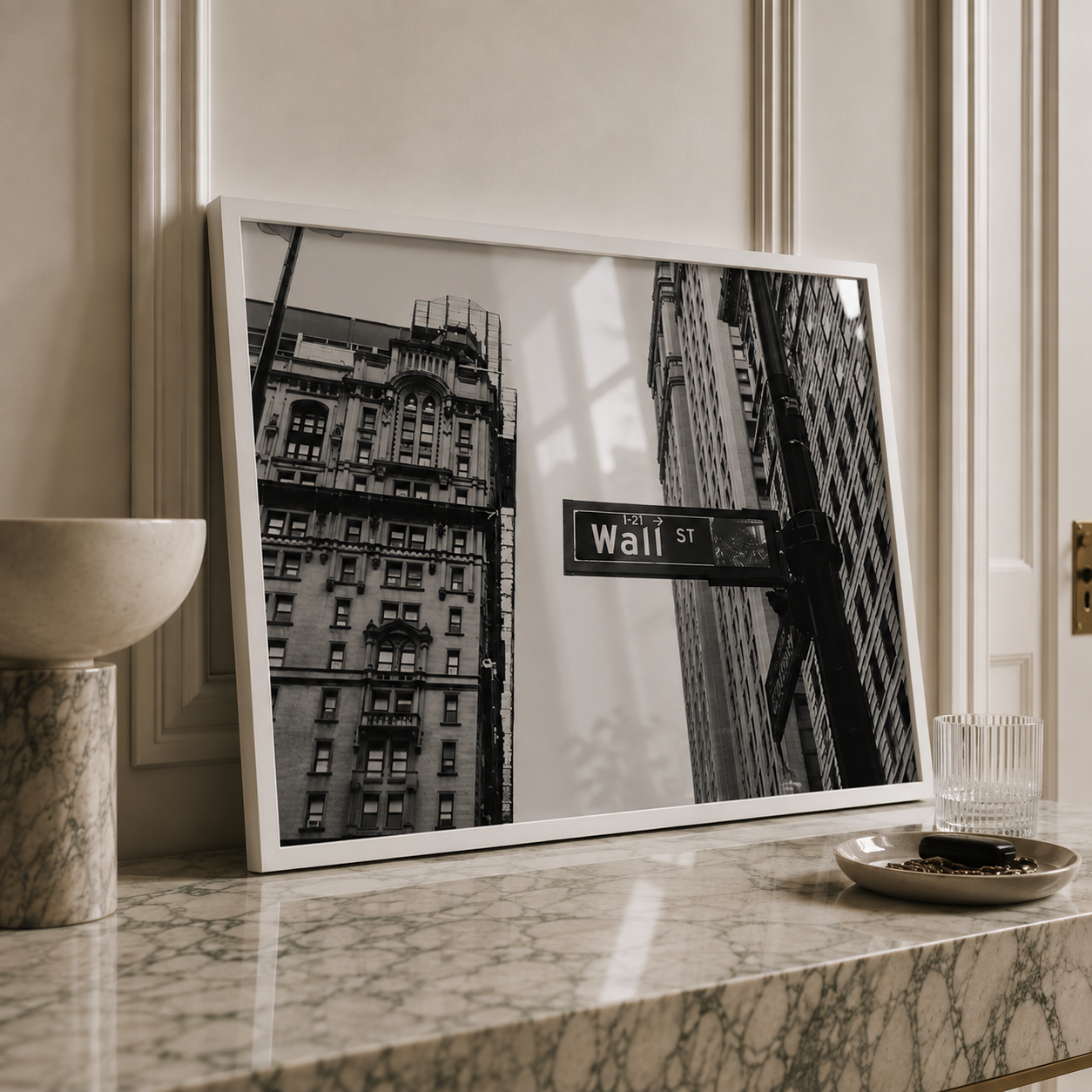 A framed black and white photograph of a New York City building facade with a visible 'Wall St' street sign, leaning against a cream paneled wall on a marble countertop with a bowl, glass, and small dish nearby.
