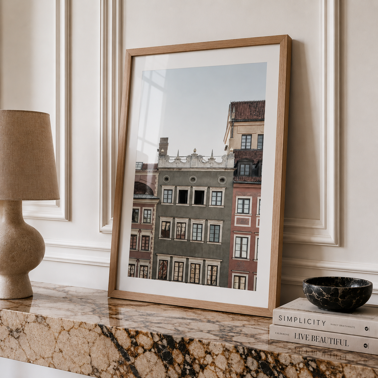 A framed art print depicting classic European townhouse facades, styled on a polished marble mantelpiece next to a lamp and books.