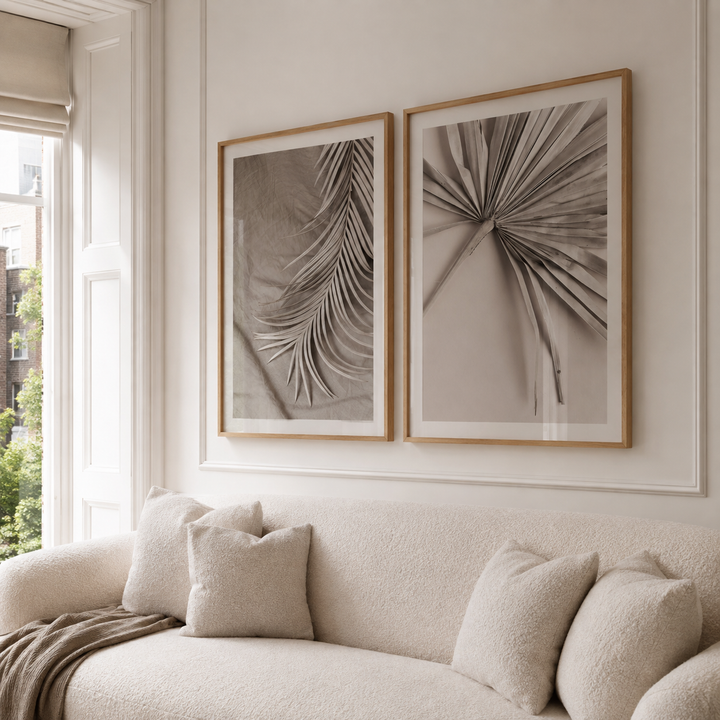 Two framed botanical art prints with light wood frames hang on a cream wall above a beige textured sofa with neutral throw pillows.