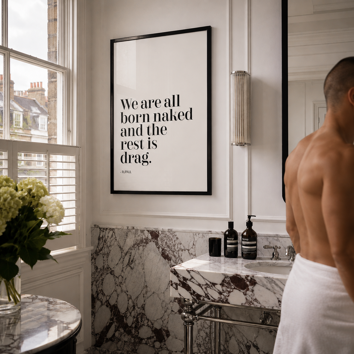 A framed black-and-white typographic wall print quote by RuPaul reading 'We are all born naked and the rest is drag.' hangs in a modern bathroom with a marble sink, marble wall paneling, a window, and a shirtless man wrapped in a white towel standing nearby.