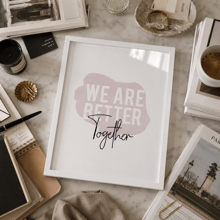 A framed typographic wall art print reading “WE ARE BETTER Together” sits on a marble surface surrounded by books, a candle, a cup of coffee, and small decorative objects. The print has a white background with a soft pink abstract shape and white block text plus black script text.