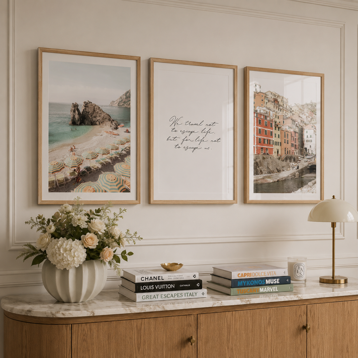 Three framed wall art prints hang above a marble console table in a styled interior. The left print shows a coastal beach scene with umbrellas and rocks; the center print has cursive script text on a white background; the right print shows colorful coastal buildings by the water.