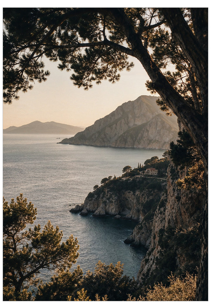 A scenic coastal landscape print showing a rocky sea cliff, calm blue water, distant mountains, and warm golden sunlight, framed by dark tree branches and foliage in the foreground.
