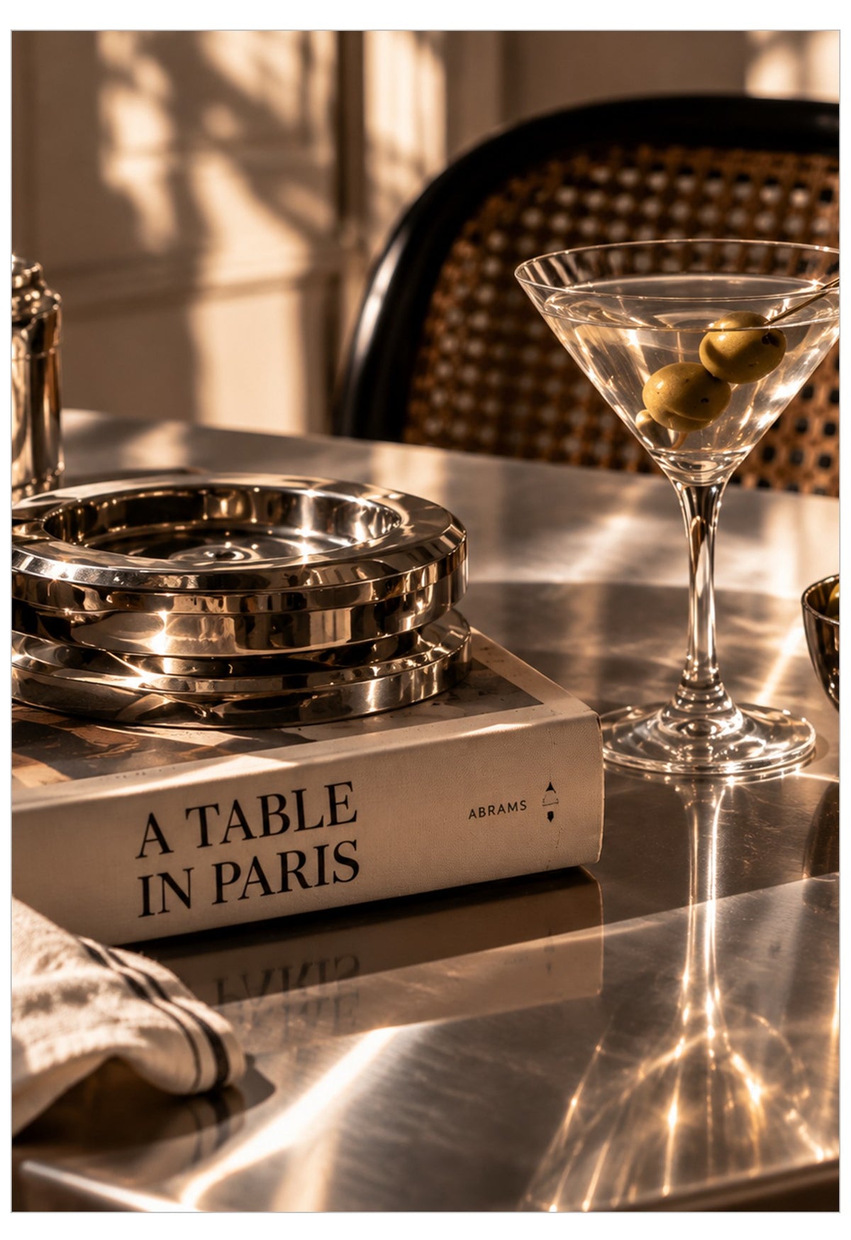 A styled tabletop scene with a martini glass containing olives, a stack of metallic trays, and a book titled "A TABLE IN PARIS" on a reflective surface in warm sunlight.