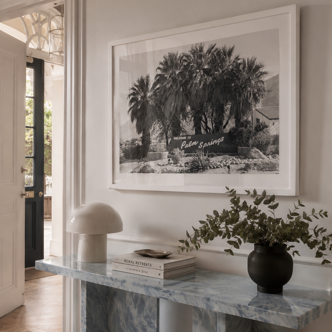 A large framed black and white art print depicting palm trees hangs on a light beige wall above a marble console table styled with a mushroom lamp, books, and a potted plant.