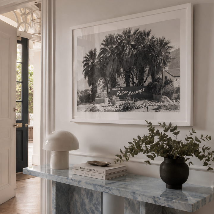 A large framed black and white art print depicting palm trees hangs on a light beige wall above a marble console table styled with a mushroom lamp, books, and a potted plant.