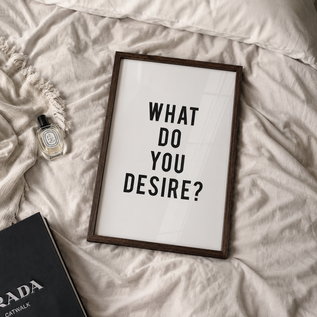 A framed typographic wall art print reading “WHAT DO YOU DESIRE?” in black text on a white background, styled on rumpled cream bedding with a small perfume bottle and part of a book nearby.