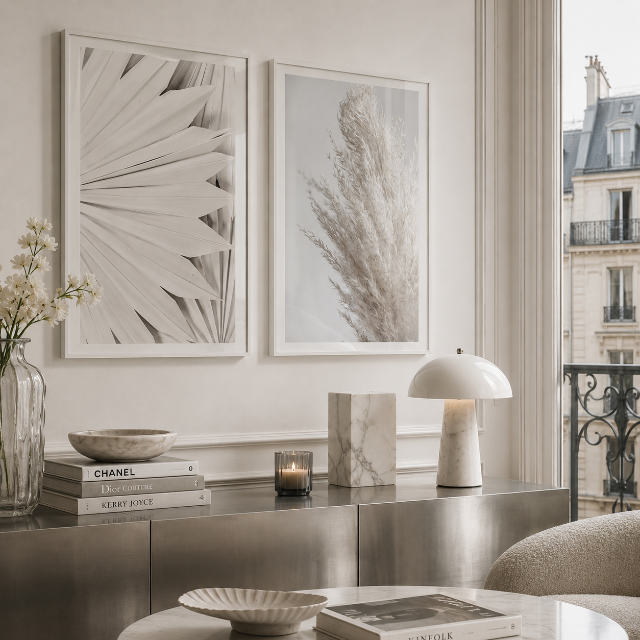 Two framed botanical wall art prints in a bright, modern living room setting. The prints feature monochrome designs: one with layered fan-like leaves and the other with pampas grass, hung side-by-side on a light beige wall above a metallic console table.