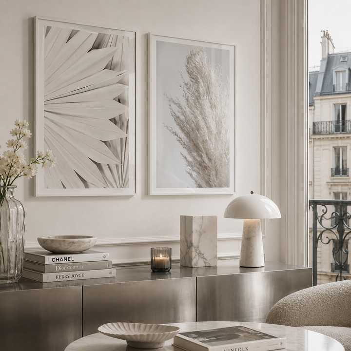 Two framed botanical wall art prints in a bright, modern living room setting. The prints feature monochrome designs: one with layered fan-like leaves and the other with pampas grass, hung side-by-side on a light beige wall above a metallic console table.