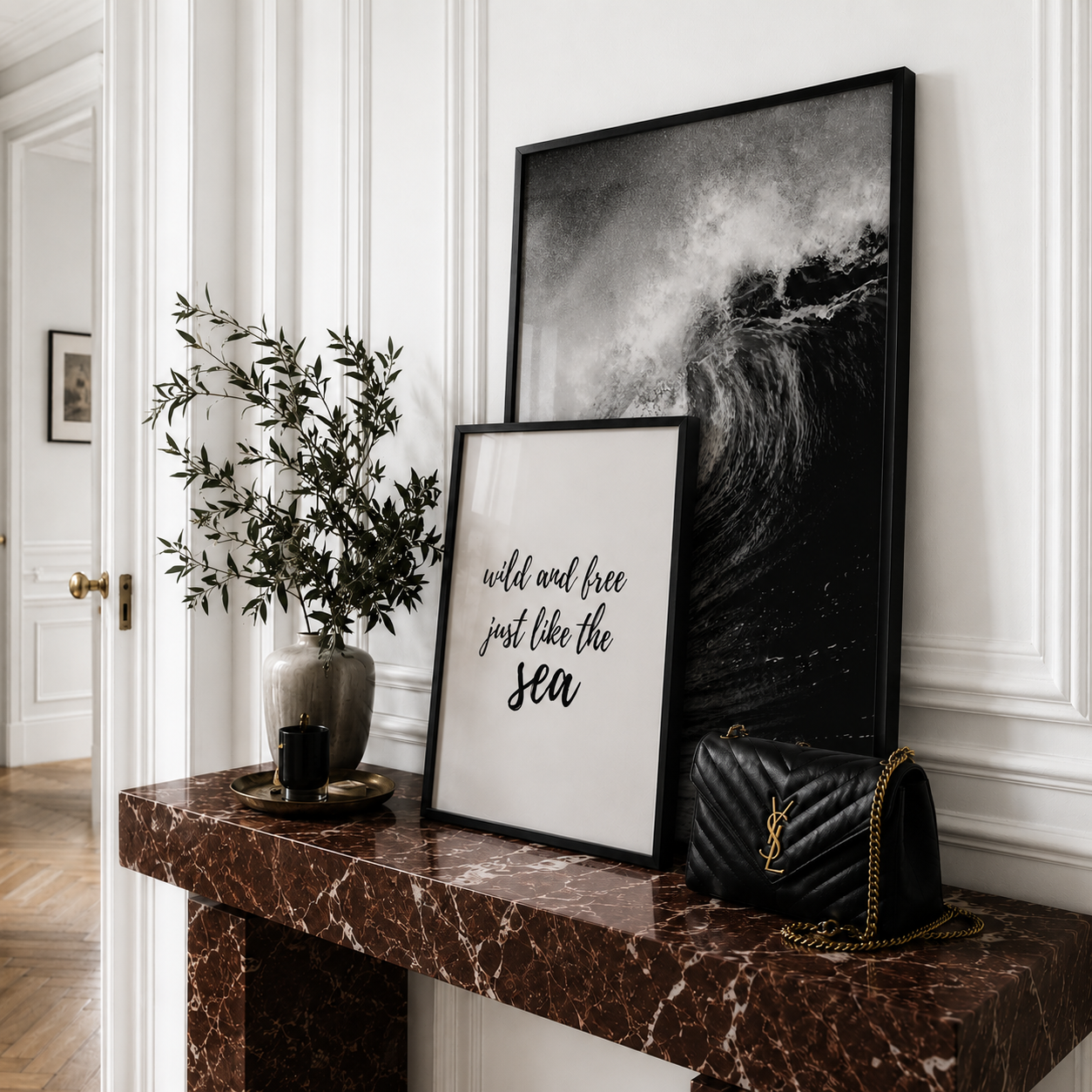 A styled interior scene with two framed wall art prints leaning on a dark marble console table. The larger print is a black-and-white ocean wave in a black frame, and the smaller print reads 'wild and free just like the sea' in black script on a white background.