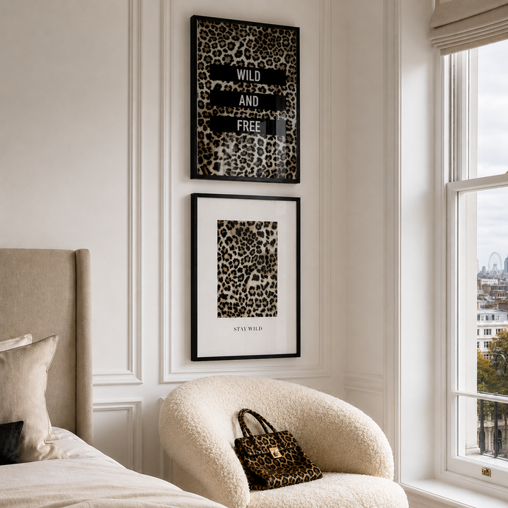 Two framed leopard print wall art prints hang on a cream paneled wall above a beige armchair in a styled bedroom. One print has black text reading 'WILD AND FREE' over a leopard pattern, and the other is a minimalist leopard print with 'WILD LIFE' text.