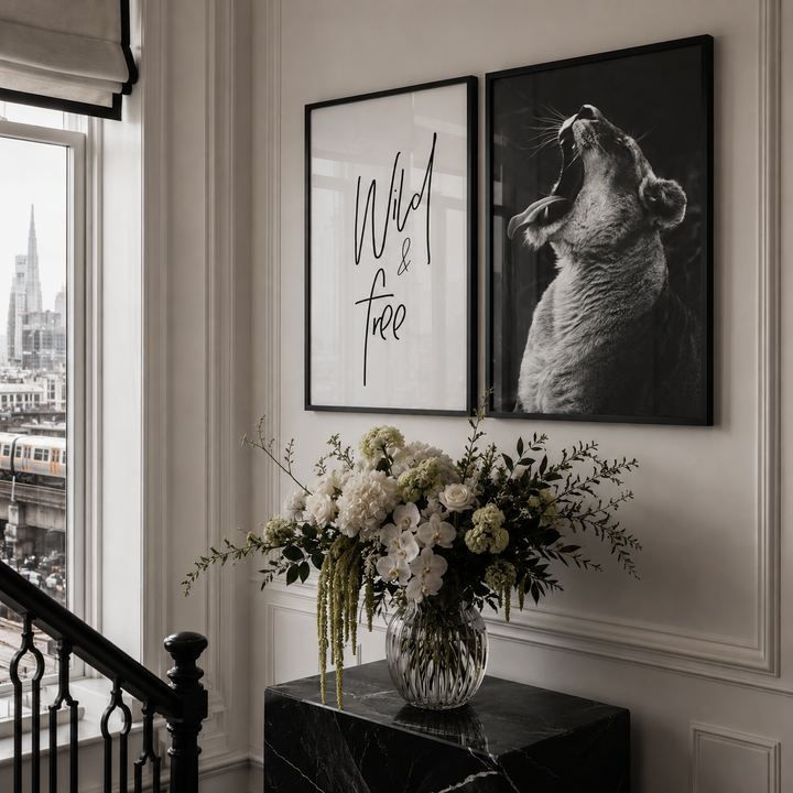 Two framed wall art prints displayed in a real interior setting: one with black script text on a white background reading 'Wild & Free,' and one black-and-white animal portrait. A floral arrangement sits on a dark marble side table beside a window and staircase railing.