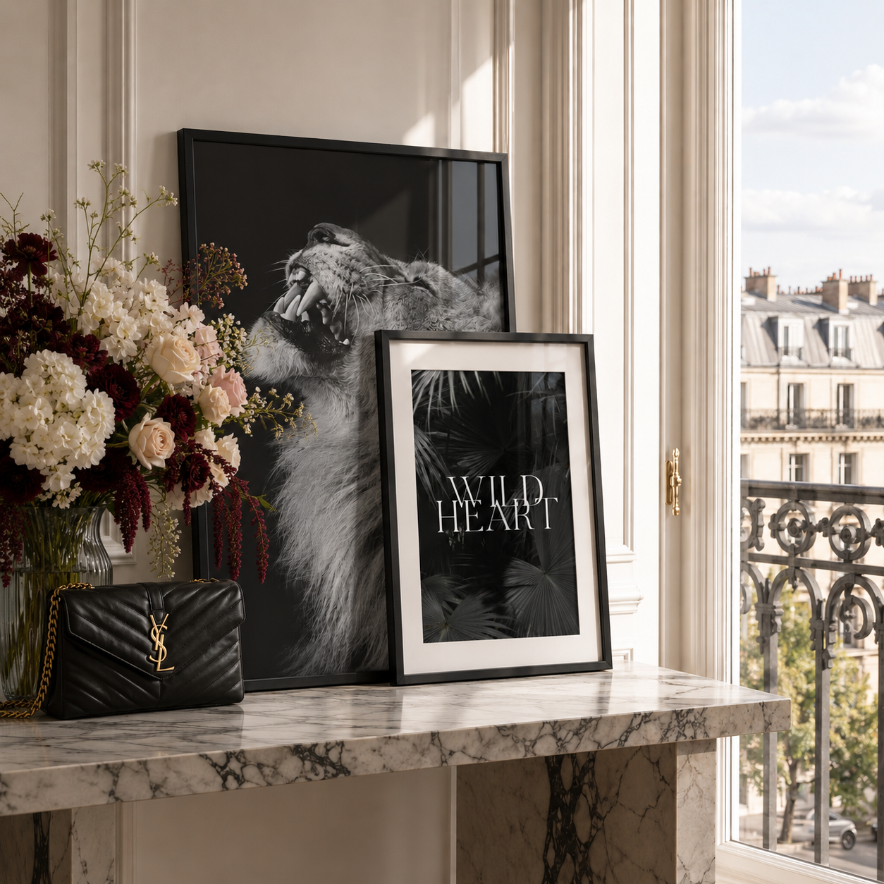 A styled interior scene with two framed black-and-white art prints leaning on a marble console table beside a black quilted YSL shoulder bag and a floral arrangement. The larger print shows a lion, and the smaller print reads “WILD HEART.