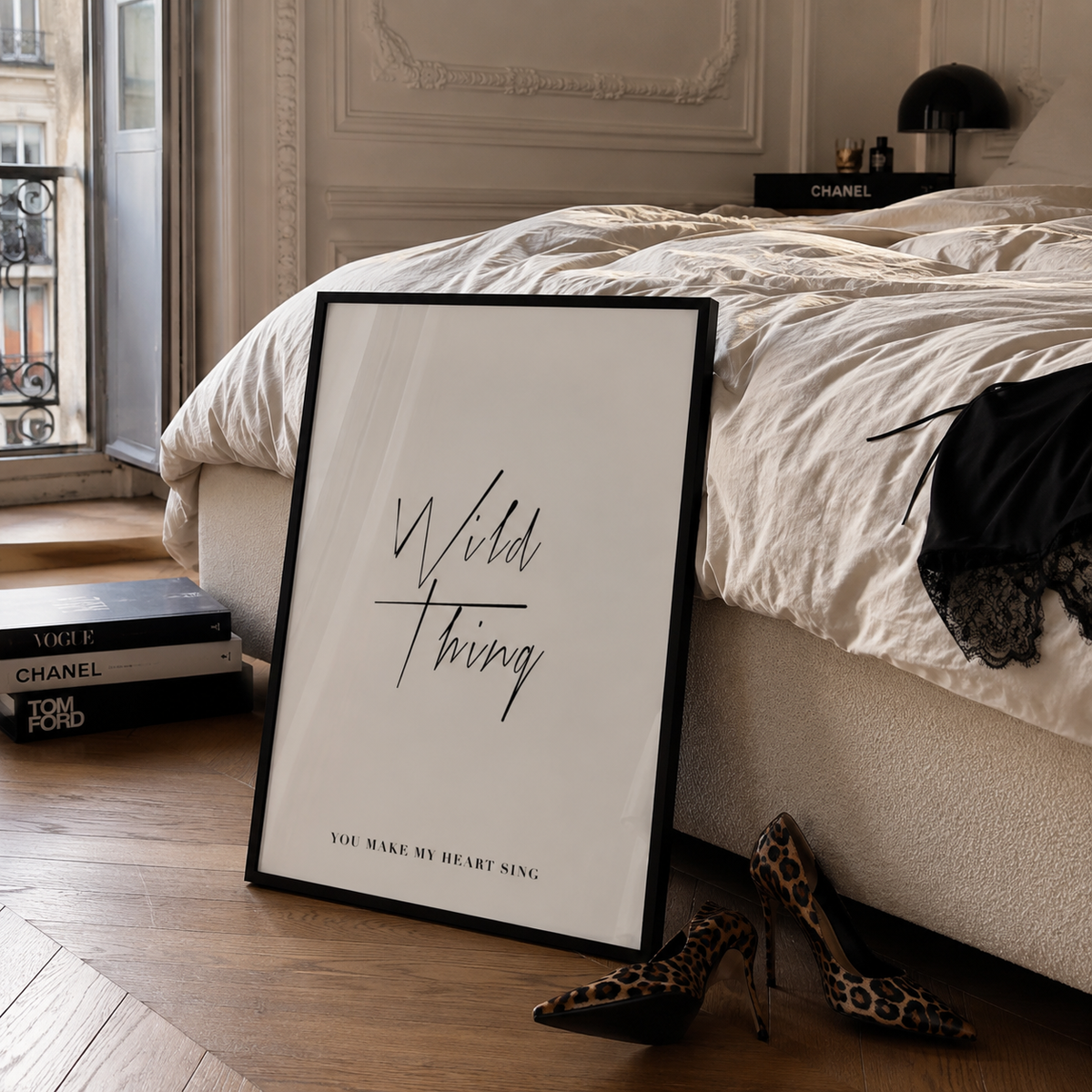 A framed black-and-white art print leaning on a wooden floor in a bedroom, with a bed, books, shoes, and a black lamp visible around it. The print reads “Wild Thing” with smaller text below.
