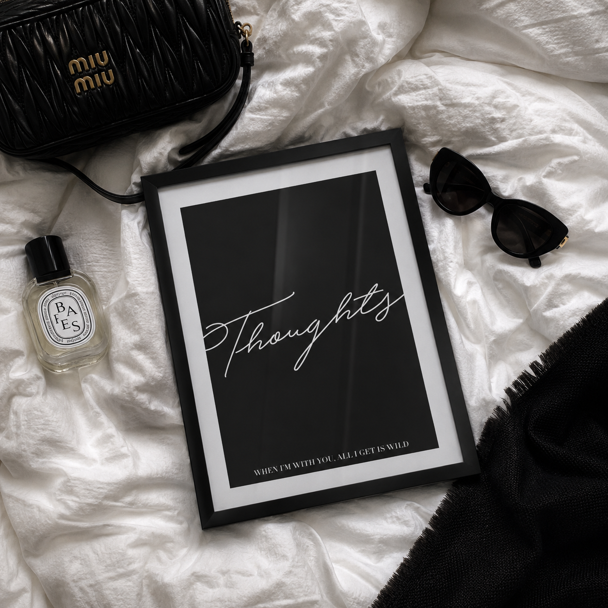 A framed black art print with the word 'Thoughts' in white script, styled in a flat lay on white fabric with a black Miu Miu bag, Diptyque perfume bottle, black sunglasses, and a black fringed textile.